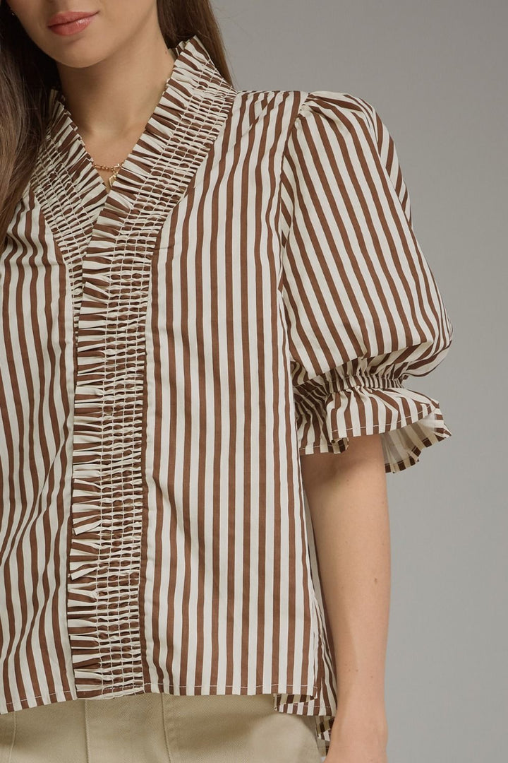 A close-up of a woman wearing a brown and beige vertically striped blouse with ruffled trim and puff sleeves, paired with light beige pants, against a neutral gray background.