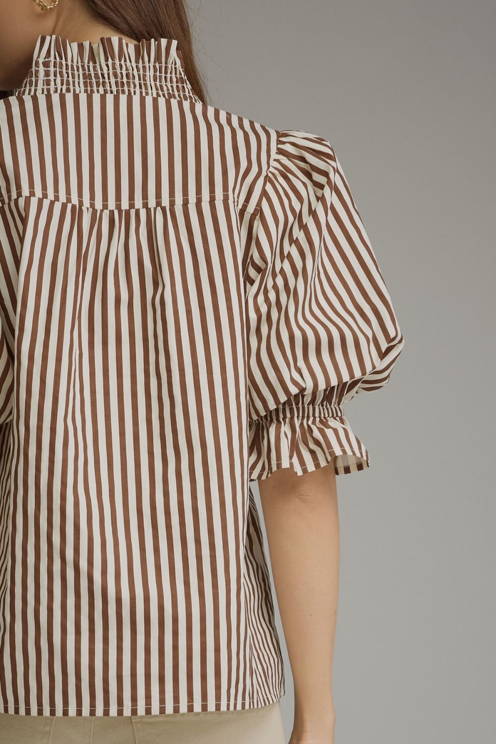 A close-up of a model wearing a brown and white vertically striped blouse with a smocked high neckline and puffed elbow-length sleeves with ruffled cuffs.