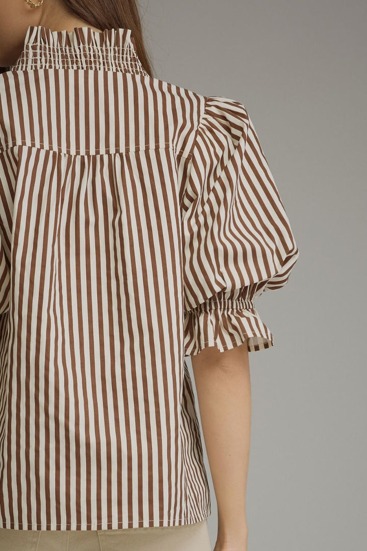 A close-up of a model wearing a brown and white vertically striped blouse with a smocked high neckline and puffed elbow-length sleeves with ruffled cuffs.