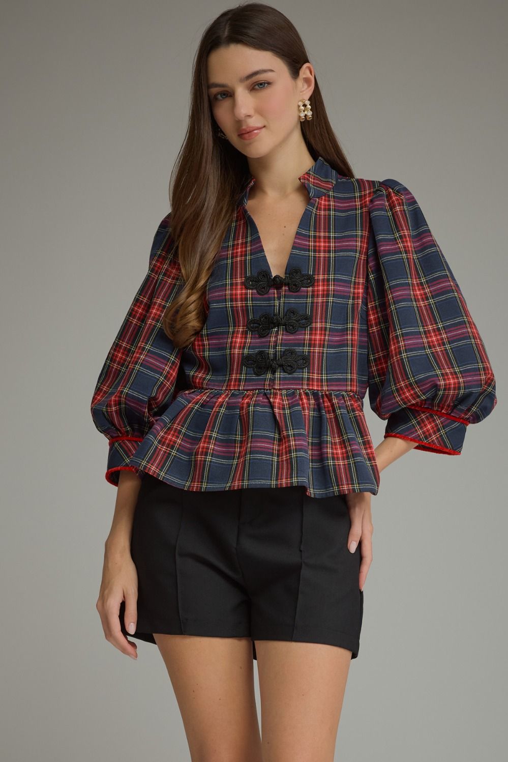 A woman modeling a navy blue plaid blouse with red and yellow stripes, featuring puff sleeves, a stand collar, and decorative black buttons down the front, paired with black shorts against a neutral gray background.