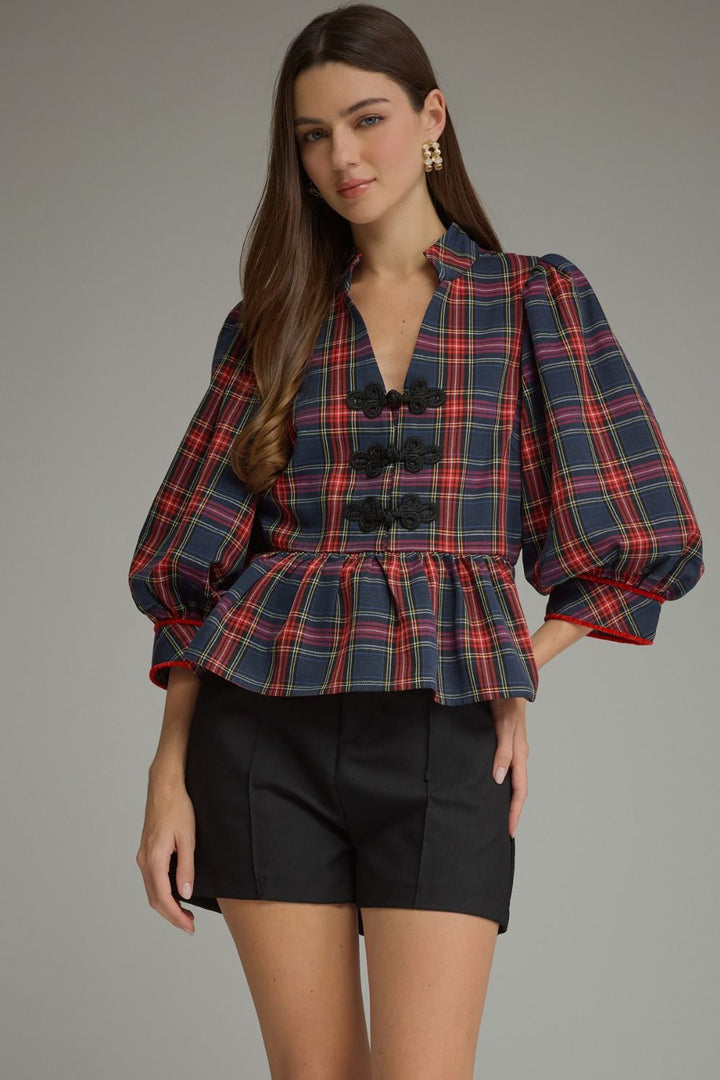 A woman modeling a navy blue plaid blouse with red and yellow stripes, featuring puff sleeves, a stand collar, and decorative black buttons down the front, paired with black shorts against a neutral gray background.