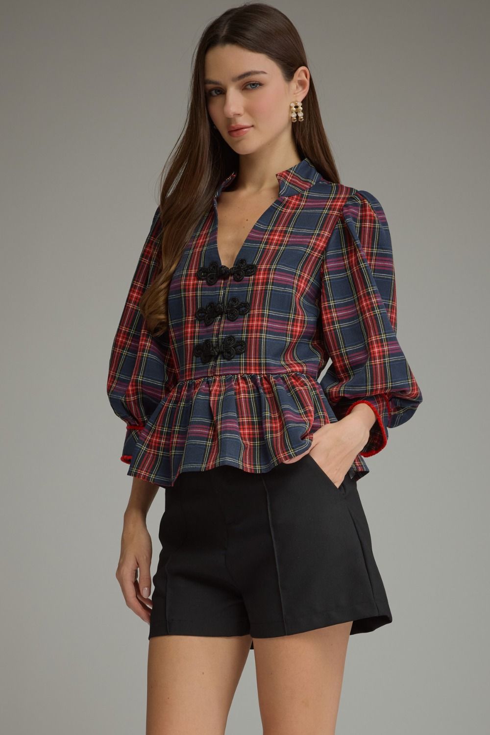 A woman modeling a navy blue plaid blouse with red and pink lines, featuring a deep V-neck, black decorative buttons, and a peplum waist, paired with black shorts against a neutral gray background.