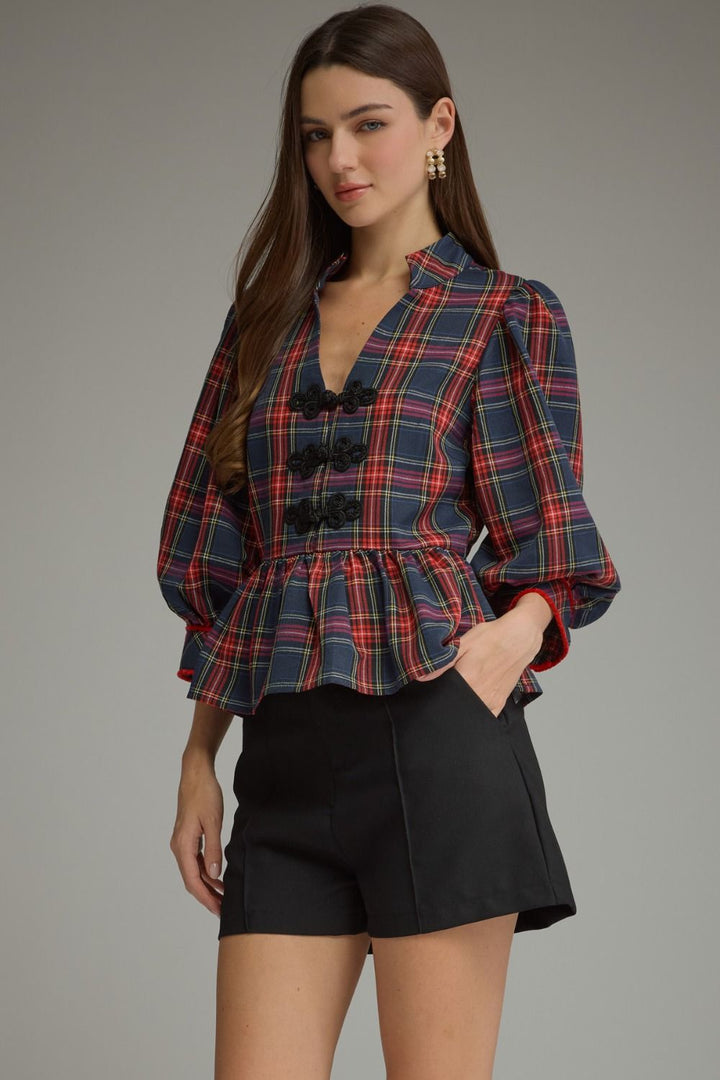 A woman modeling a navy blue plaid blouse with red and pink lines, featuring a deep V-neck, black decorative buttons, and a peplum waist, paired with black shorts against a neutral gray background.