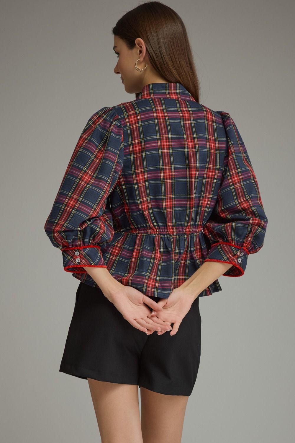 A woman shown from the back wearing a navy blue plaid shirt with red and pink lines, featuring puff sleeves and a gathered waist, paired with black shorts against a solid gray studio background.
