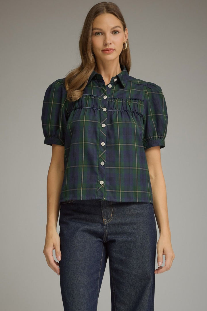 A woman modeling a short-sleeve button-up blouse with puff sleeves and a green and navy plaid pattern, paired with dark blue jeans, against a solid gray studio background.