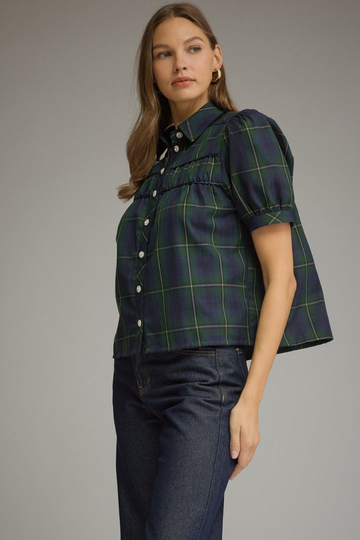 A woman modeling a short-sleeve button-front plaid blouse with puff sleeves in dark green and navy, paired with dark blue jeans, against a plain gray studio background.