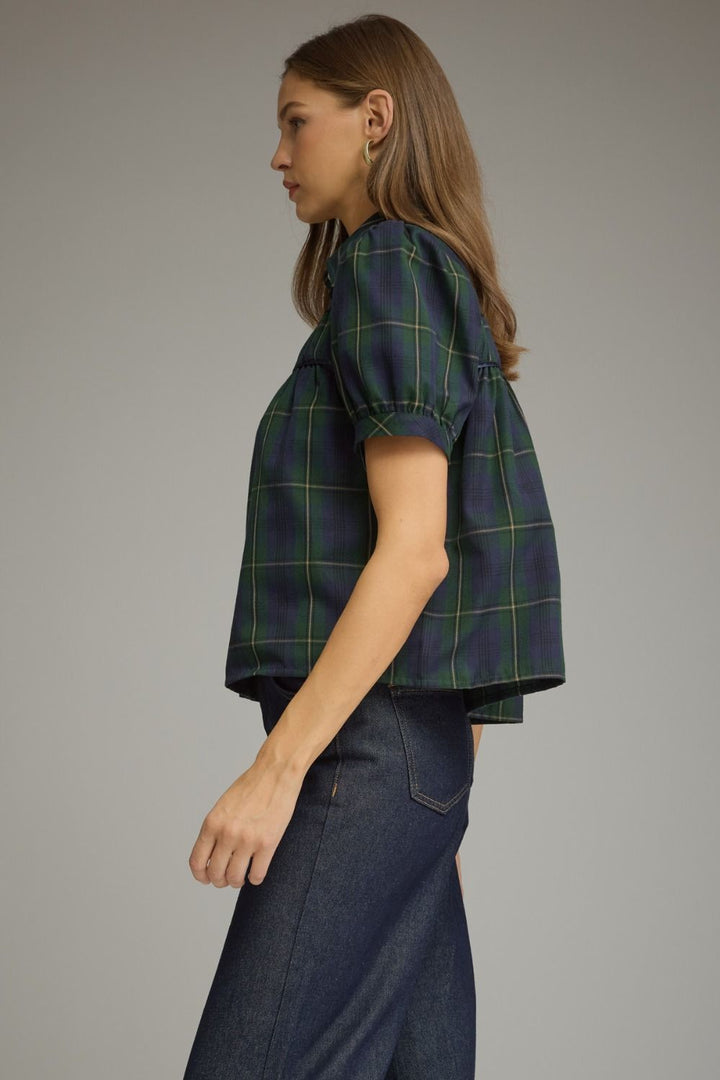 A woman shown in side profile wearing a dark navy cropped short-sleeve top with a green and yellow windowpane plaid pattern, paired with dark blue jeans, against a solid gray studio background.