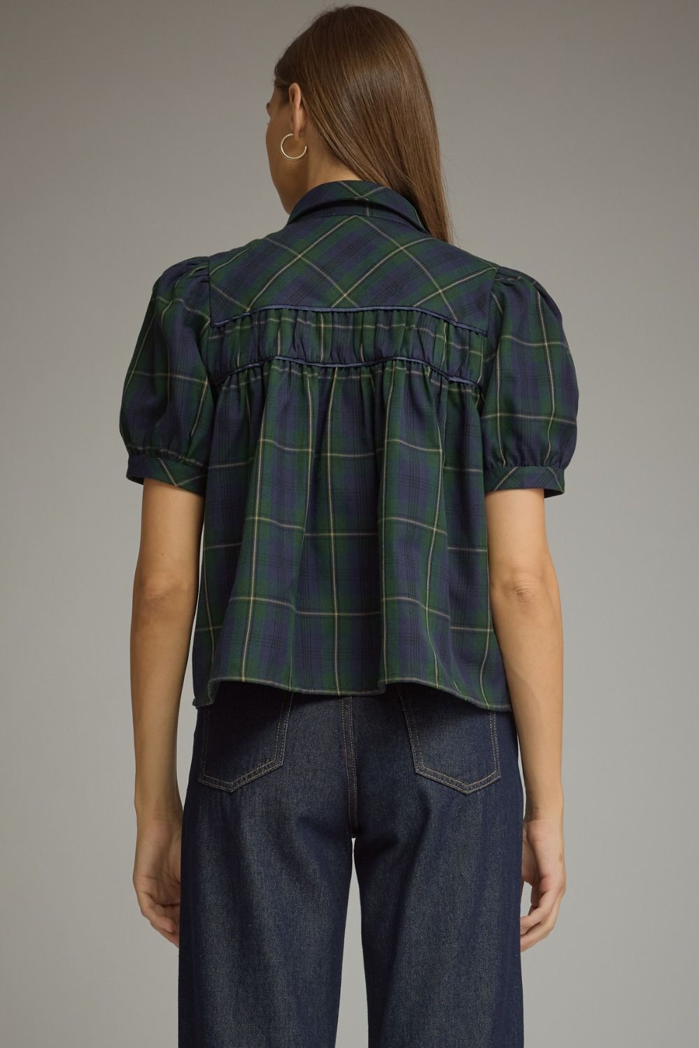 A woman shown from the back wearing a dark navy plaid short-sleeve blouse with puff sleeves and a gathered waist, paired with dark blue jeans, against a solid gray studio background.