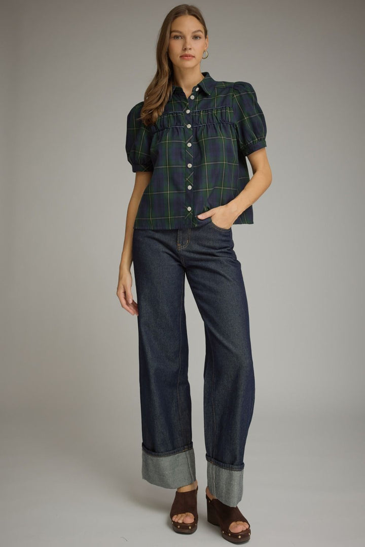 A model stands against a neutral gray studio background wearing a dark green and black plaid puff-sleeve button-up shirt, dark blue wide-leg jeans with rolled cuffs, and brown wedge sandals.
