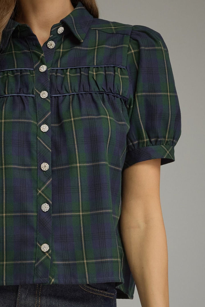 A close-up of a woman wearing a short-sleeve plaid button-up shirt in dark green and navy with thin yellow lines, featuring silver buttons and a ruffled chest detail.