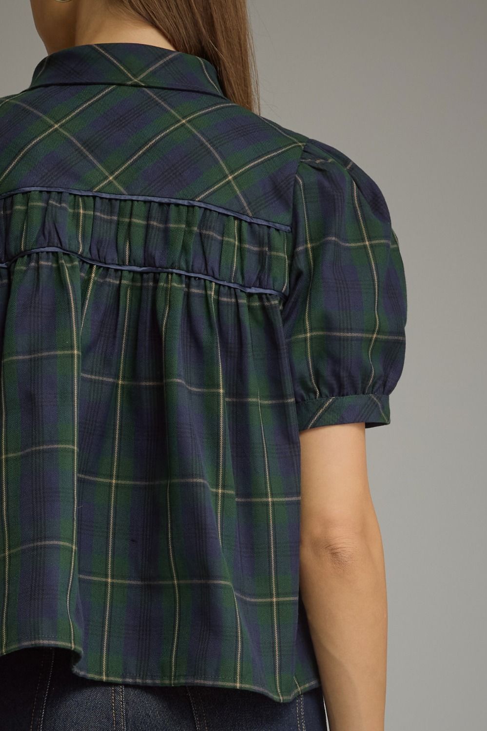 Close-up back view of a woman wearing a short-sleeve plaid blouse in dark green and navy with thin yellow lines, paired with dark jeans against a plain gray studio background.