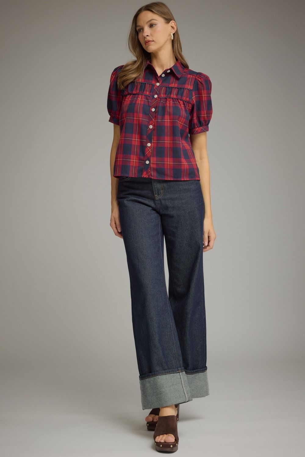 A woman modeling a short-sleeve button-front plaid shirt in red and navy with dark blue wide-leg jeans and brown sandals, standing against a plain light gray studio background.