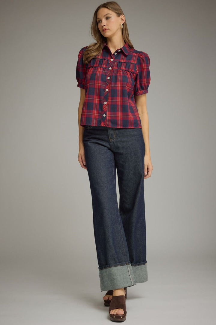 A woman modeling a short-sleeve button-front plaid shirt in red and navy with dark blue wide-leg jeans and brown sandals, standing against a plain light gray studio background.