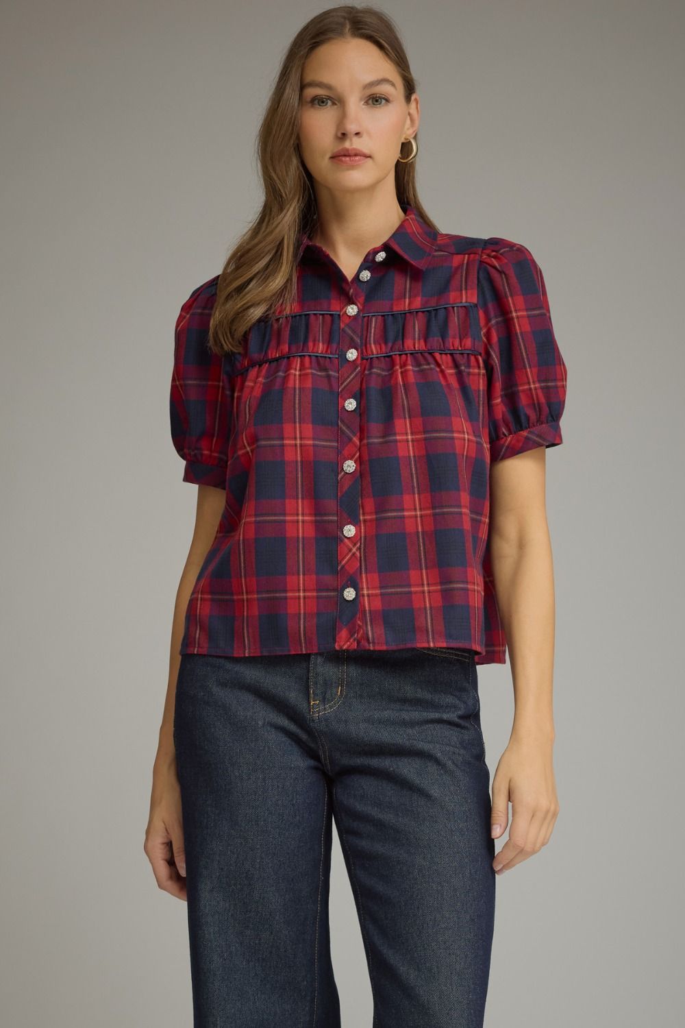 A woman modeling a short-sleeve plaid button-up shirt with puff sleeves in red and navy, paired with dark blue jeans, against a solid gray studio background.