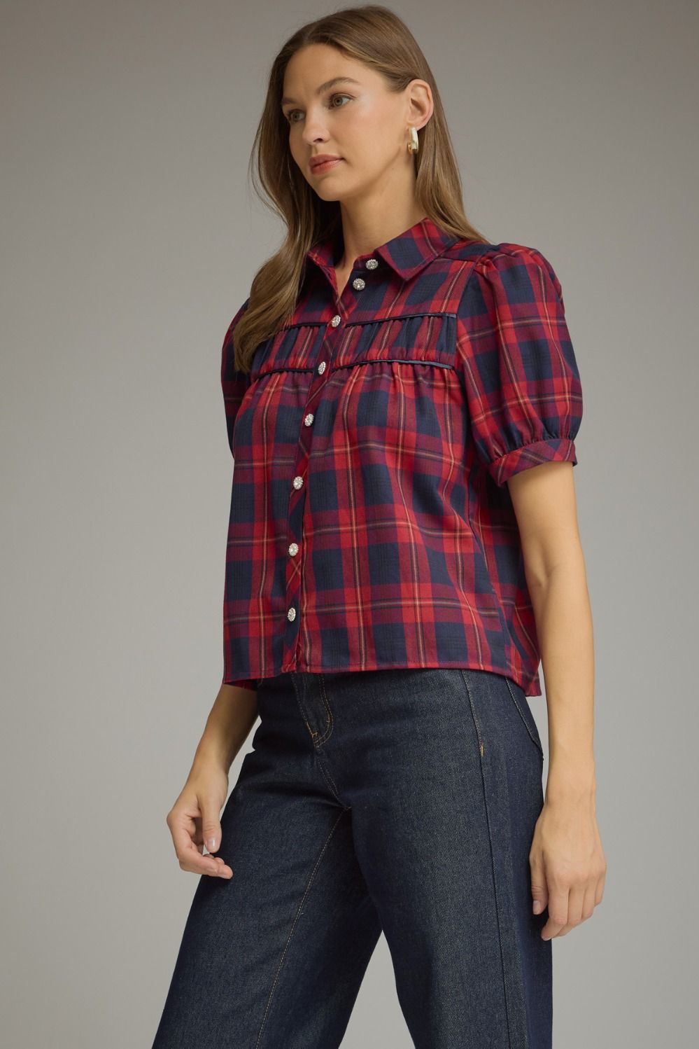 A woman modeling a short-sleeve button-front plaid blouse in red and navy, paired with dark blue jeans, against a plain gray studio background.