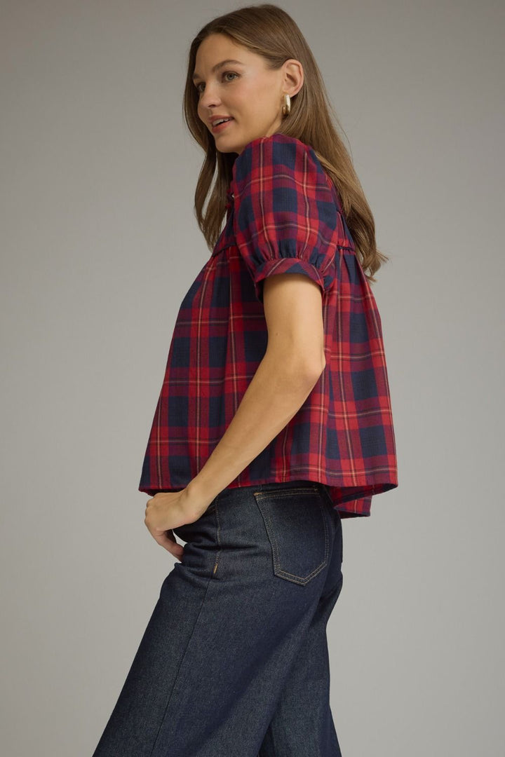 A woman wearing a red and navy blue plaid puff-sleeve blouse with dark blue jeans, photographed against a solid gray studio background.