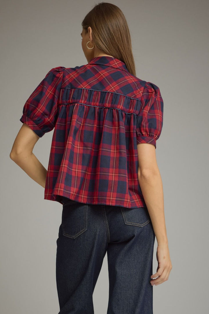 A woman shown from the back wearing a red and navy plaid puff-sleeve blouse tucked into dark blue jeans, against a solid gray studio background.