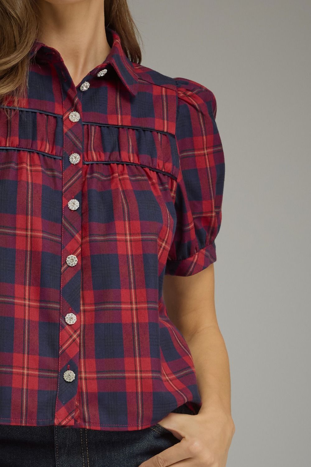 A close-up of a woman wearing a short-sleeve plaid button-up shirt with puff sleeves and silver buttons, paired with dark denim jeans against a solid gray background.