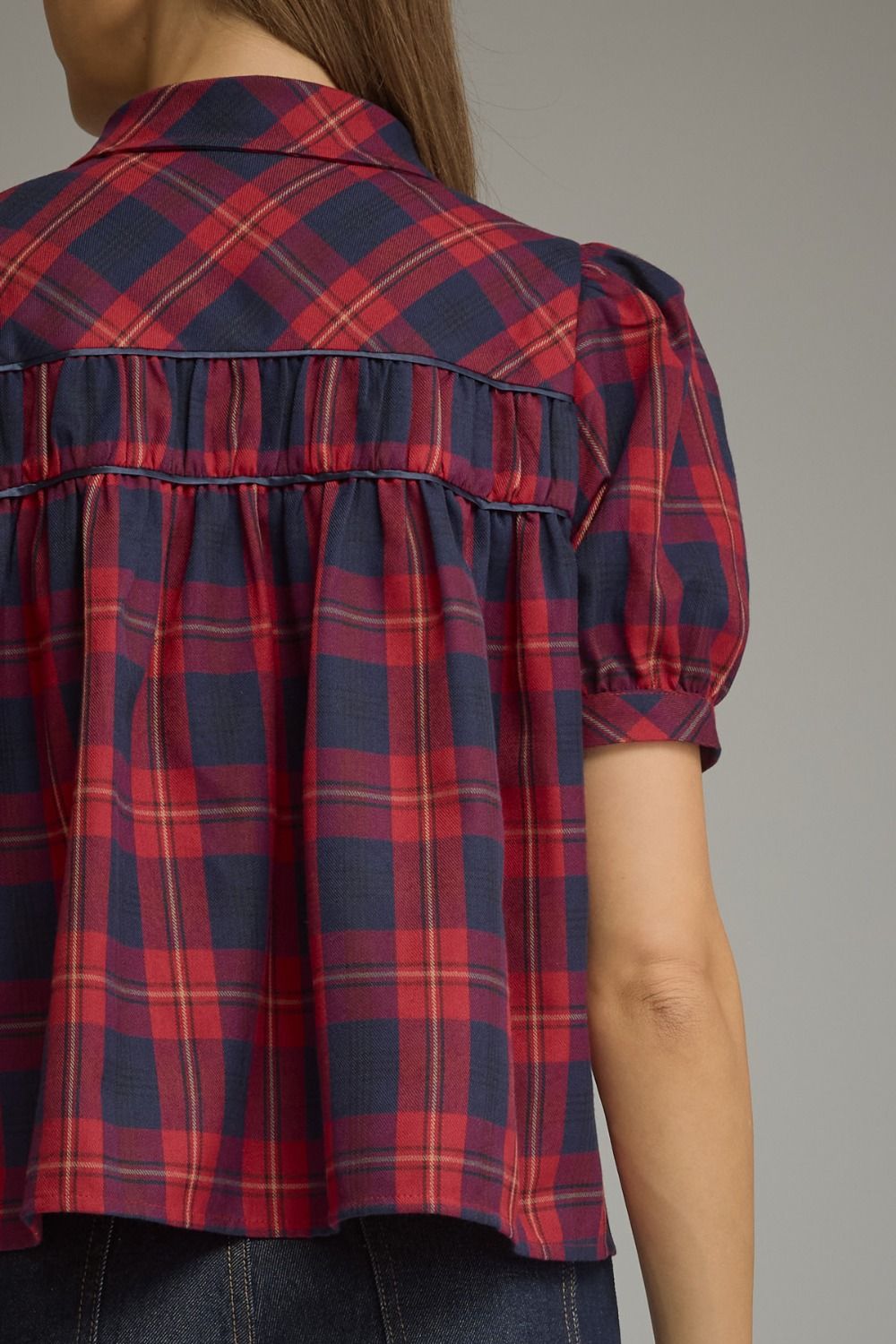 Close-up back view of a woman wearing a short-sleeve plaid blouse in navy blue, red, and thin yellow lines, paired with dark jeans against a plain gray studio background.
