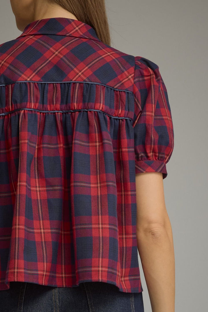Close-up back view of a woman wearing a short-sleeve plaid blouse in navy blue, red, and thin yellow lines, paired with dark jeans against a plain gray studio background.