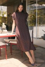 A woman modeling a dark brown midi dress with a white polka dot pattern, standing on a patio next to a table.