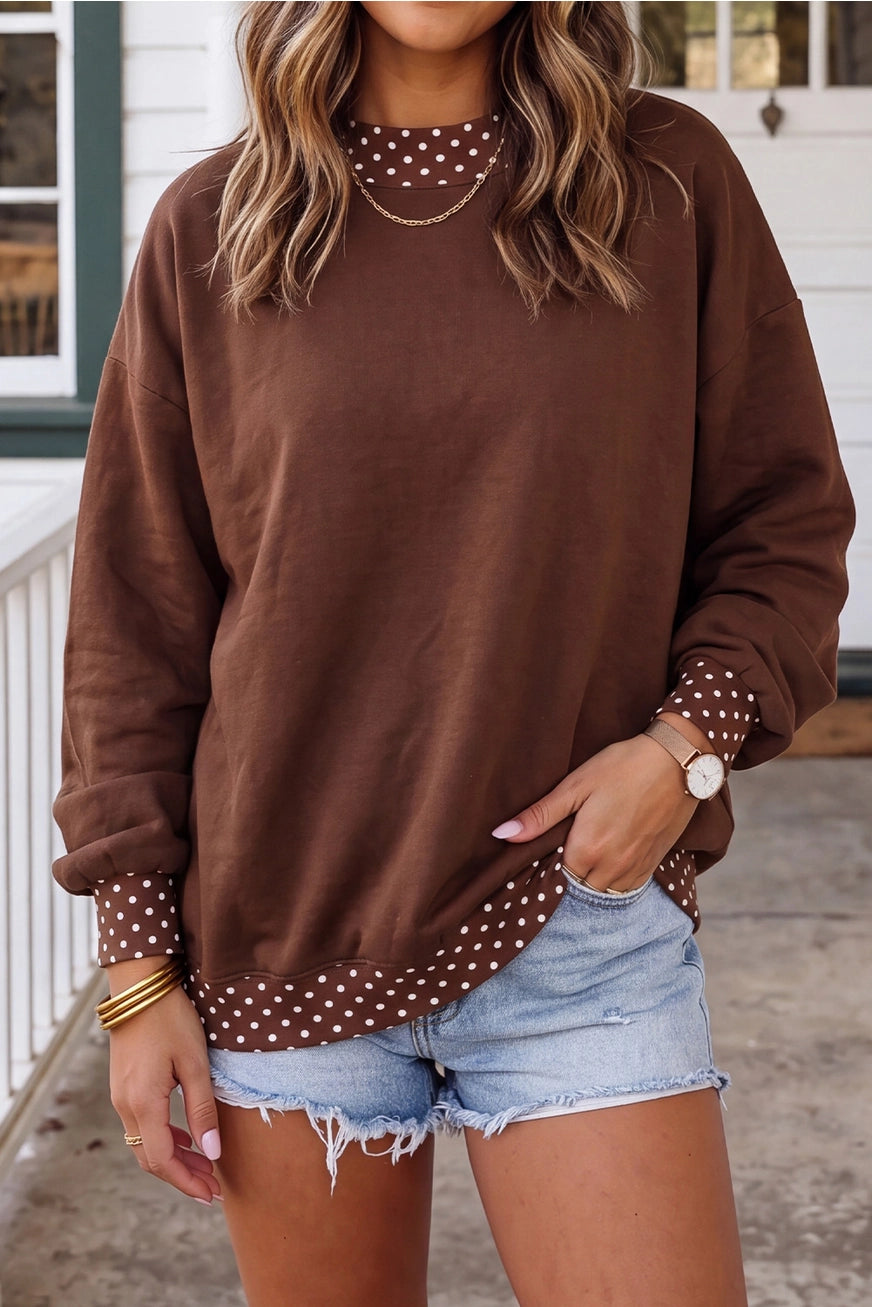 A woman wearing a brown crewneck sweatshirt with white polka dot trim along the collar, cuffs, and hem, paired with light-wash denim shorts.