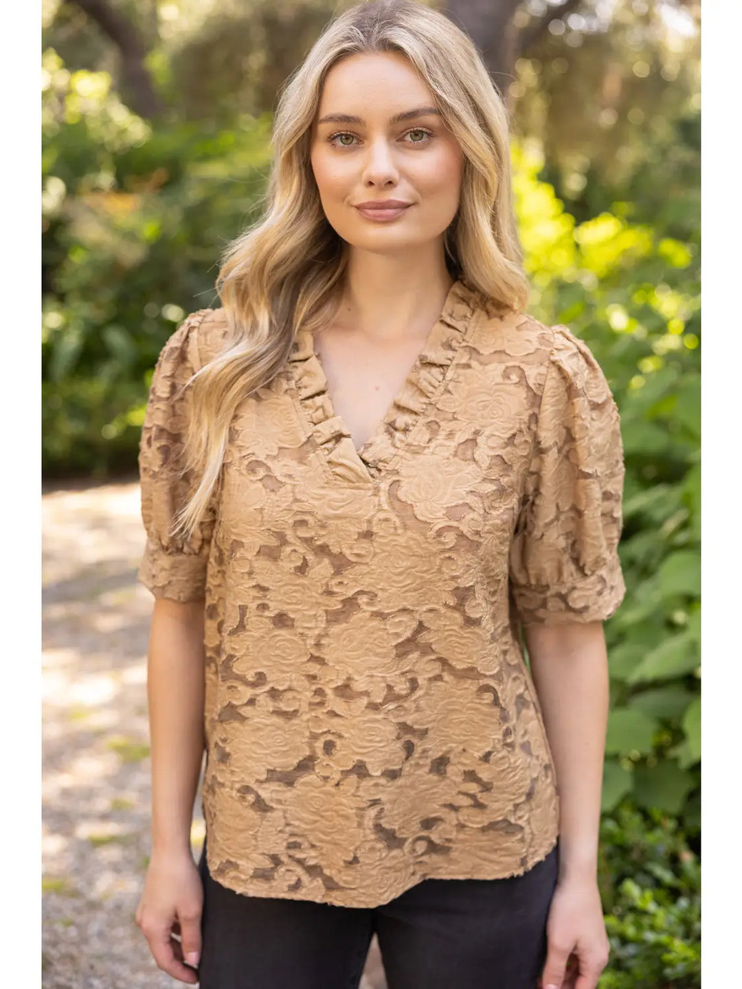 A woman wearing a tan lace short-sleeve blouse with puff sleeves and a ruffled V-neckline, standing outdoors against a blurred green garden background.
