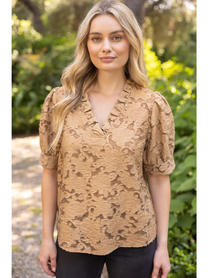 A woman wearing a tan lace short-sleeve blouse with puff sleeves and a ruffled V-neckline, standing outdoors against a blurred green garden background.