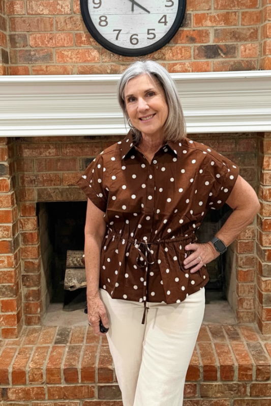 A woman with gray hair stands in front of a brick fireplace wearing a brown short-sleeve button-up shirt with white polka dots and cream pants.