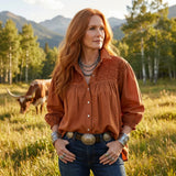 Woman in an orange shirt and blue jeans standing in a field with mountains and trees in the background.