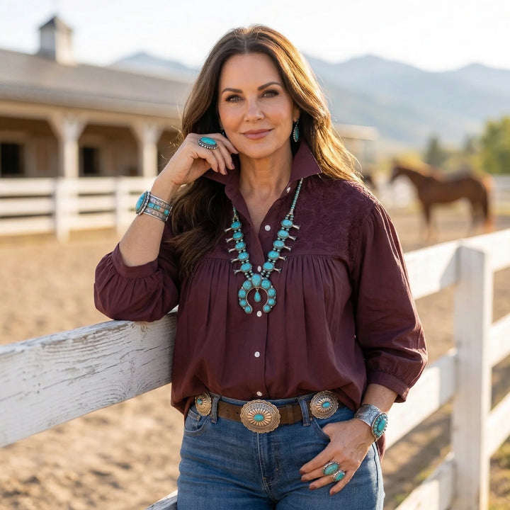 Woman in a burgundy shirt with turquoise jewelry standing by a wooden fence with a horse in the background.