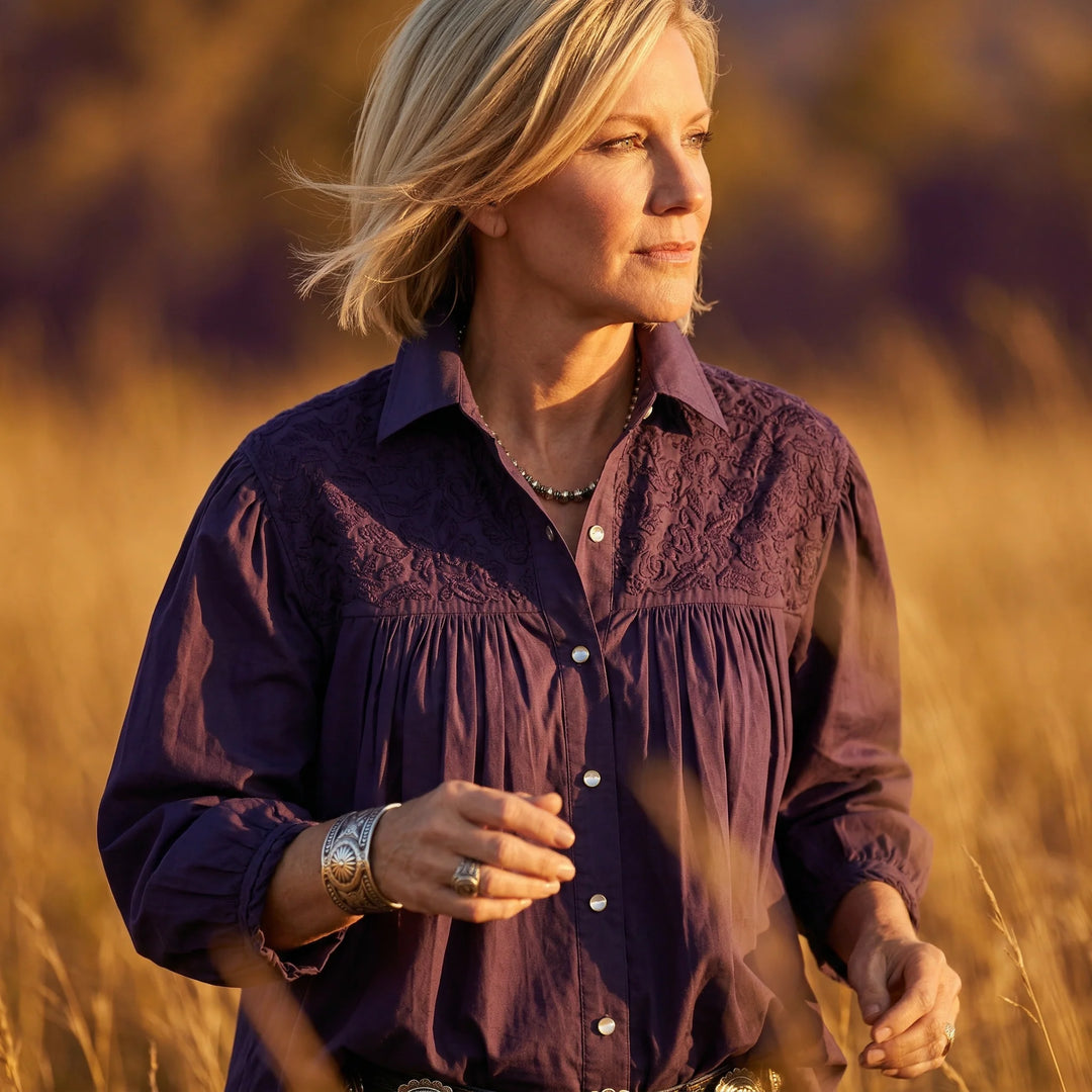 Woman in a burgundy shirt with turquoise jewelry standing by a wooden fence with a horse in the background.