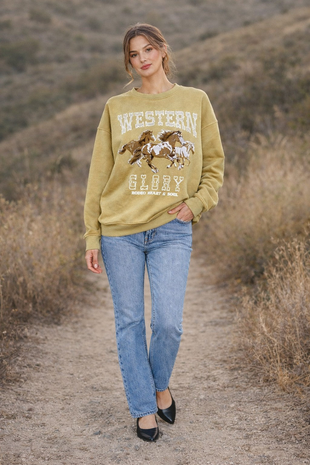 Woman wearing a mustard yellow sweatshirt with a horse graphic and text, standing on a dirt path in a natural setting.