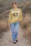 Woman wearing a mustard yellow sweatshirt with a horse graphic and text, standing on a dirt path in a natural setting.